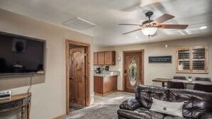 A living room with a leather couch, a flat-screen TV, a ceiling fan, and a kitchen area with wooden cabinets and a bar. - Mountain Flo by Ruidoso Vacation Rentals (Alto)