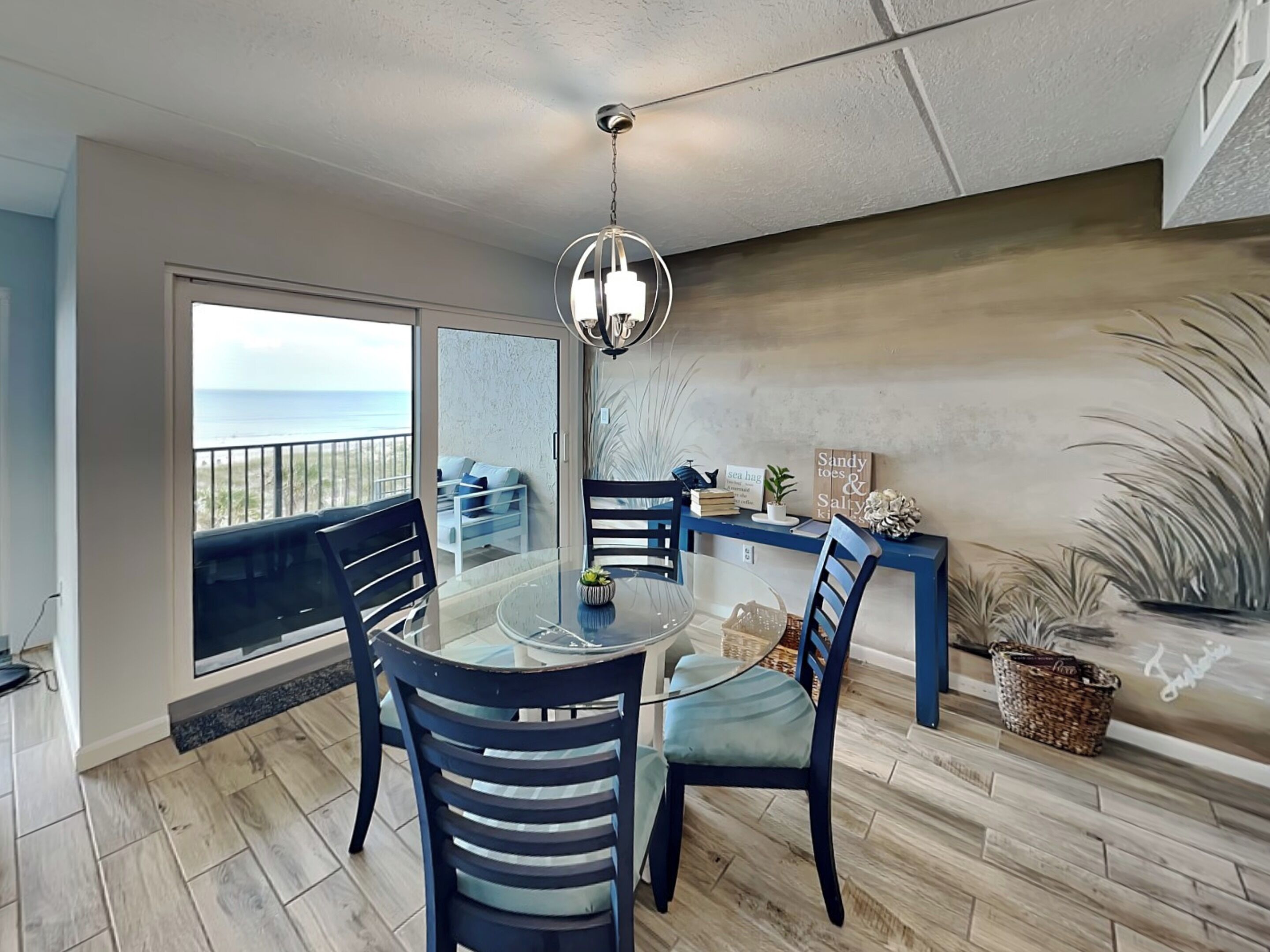 A dining area with a glass table, four chairs, a hanging light fixture, a view of the ocean, and a balcony with a sofa.