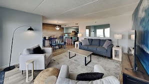 A modern living room with a grey sofa, a white armchair, and a coffee table. There is a dining area with a table and chairs, and a kitchen in the background. - Beachcomber 403 by Distinctive Beach Rentals (Jacksonville Beach)