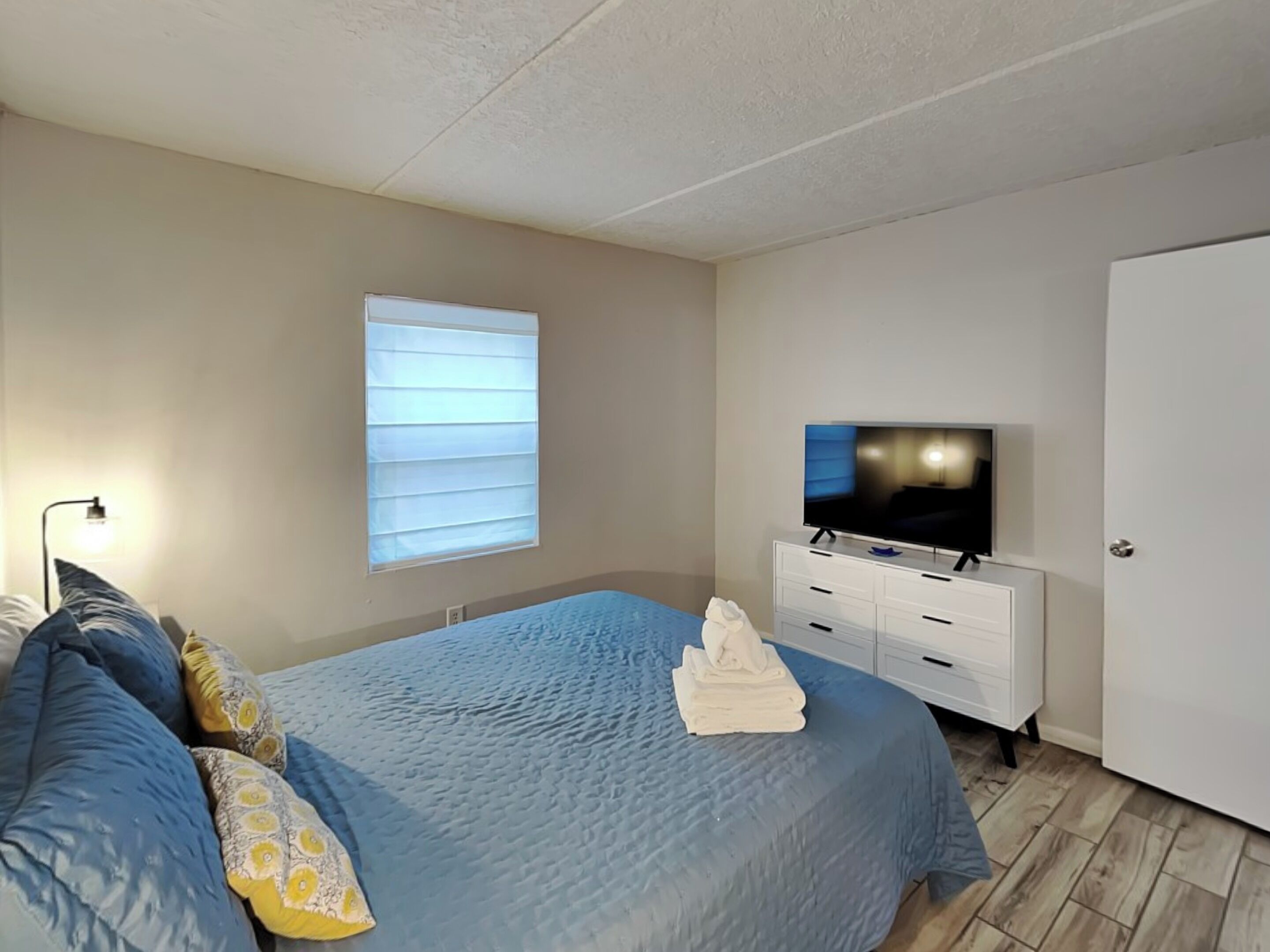 Une chambre à coucher avec un lit, un téléviseur posé sur une commode, une fenêtre avec des stores et une porte.
