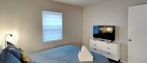 Une chambre à coucher avec un lit, un téléviseur posé sur une commode, une fenêtre avec des stores et une porte.