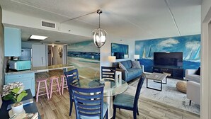 A modern living room with a glass dining table, blue chairs, a sofa, a TV, and a mural of sailboats. - Beachcomber 403 by Distinctive Beach Rentals (Jacksonville Beach)