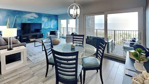 A modern living room with a round glass table, dark blue chairs, a sofa, a TV, and a large blue mural. - Beachcomber 403 by Distinctive Beach Rentals (Jacksonville Beach)