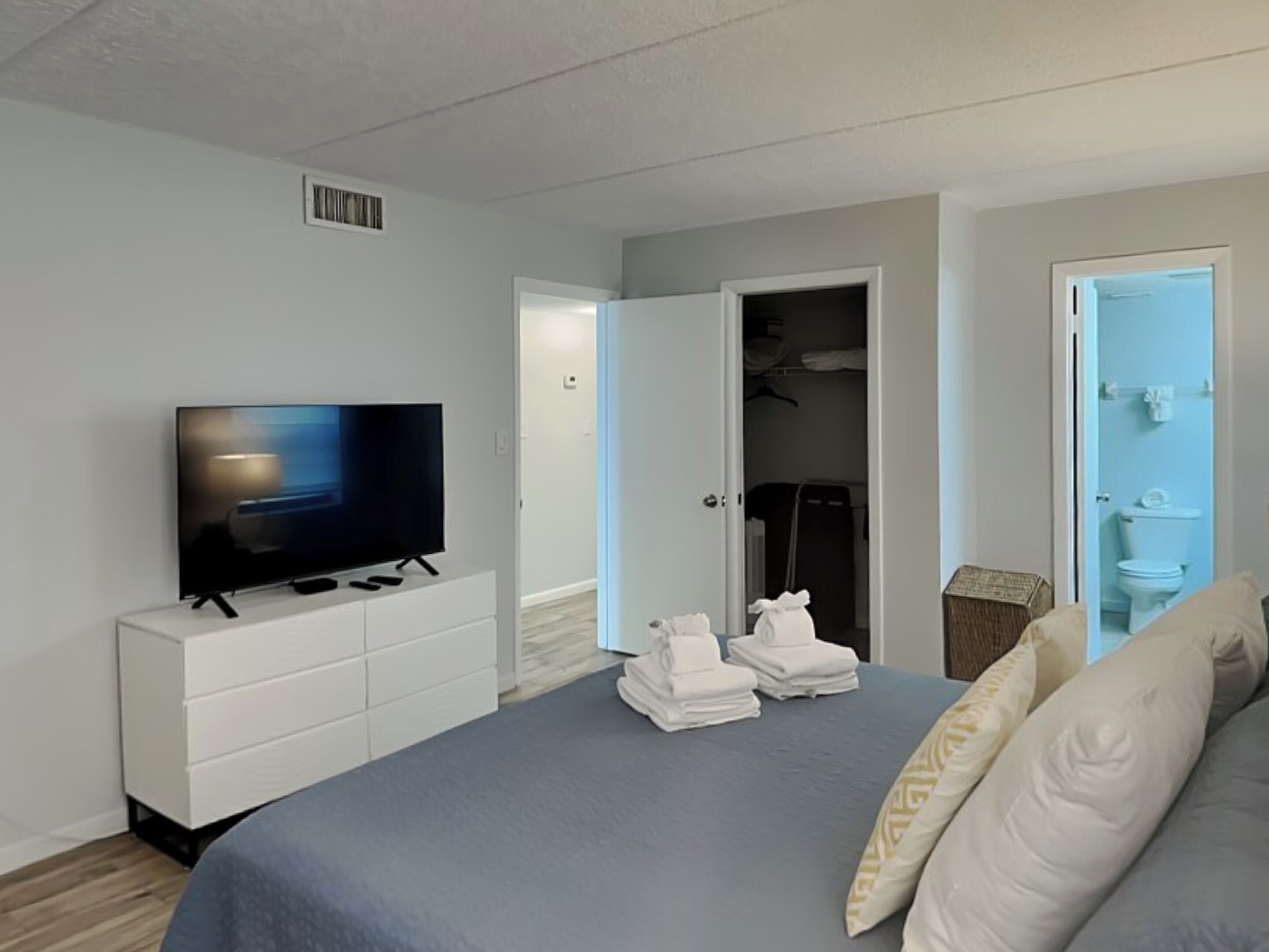 A modern hotel room with a flat-screen TV on a white dresser, a neatly made bed with pillows, a closet with hanging clothes, and a bathroom with a toilet and sink.