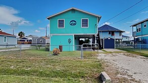 Una casa turquesa con porche cubierto, rodeada por una cerca y otras viviendas.