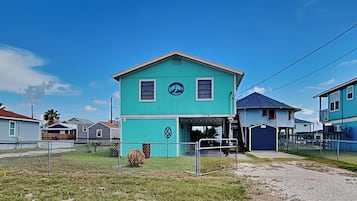 A turquoise house with a covered porch, surrounded by a fence and other houses.