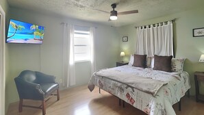 Een slaapkamer met een bed, een stoel, een plafondventilator, een televisie en een raam met gordijnen.