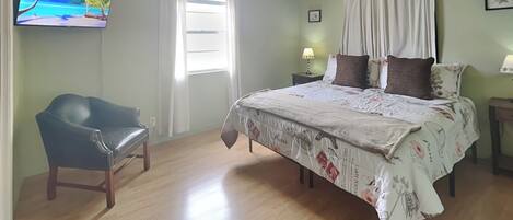 Une chambre à coucher avec un lit, une chaise, un ventilateur de plafond, une télévision et une fenêtre avec des rideaux.