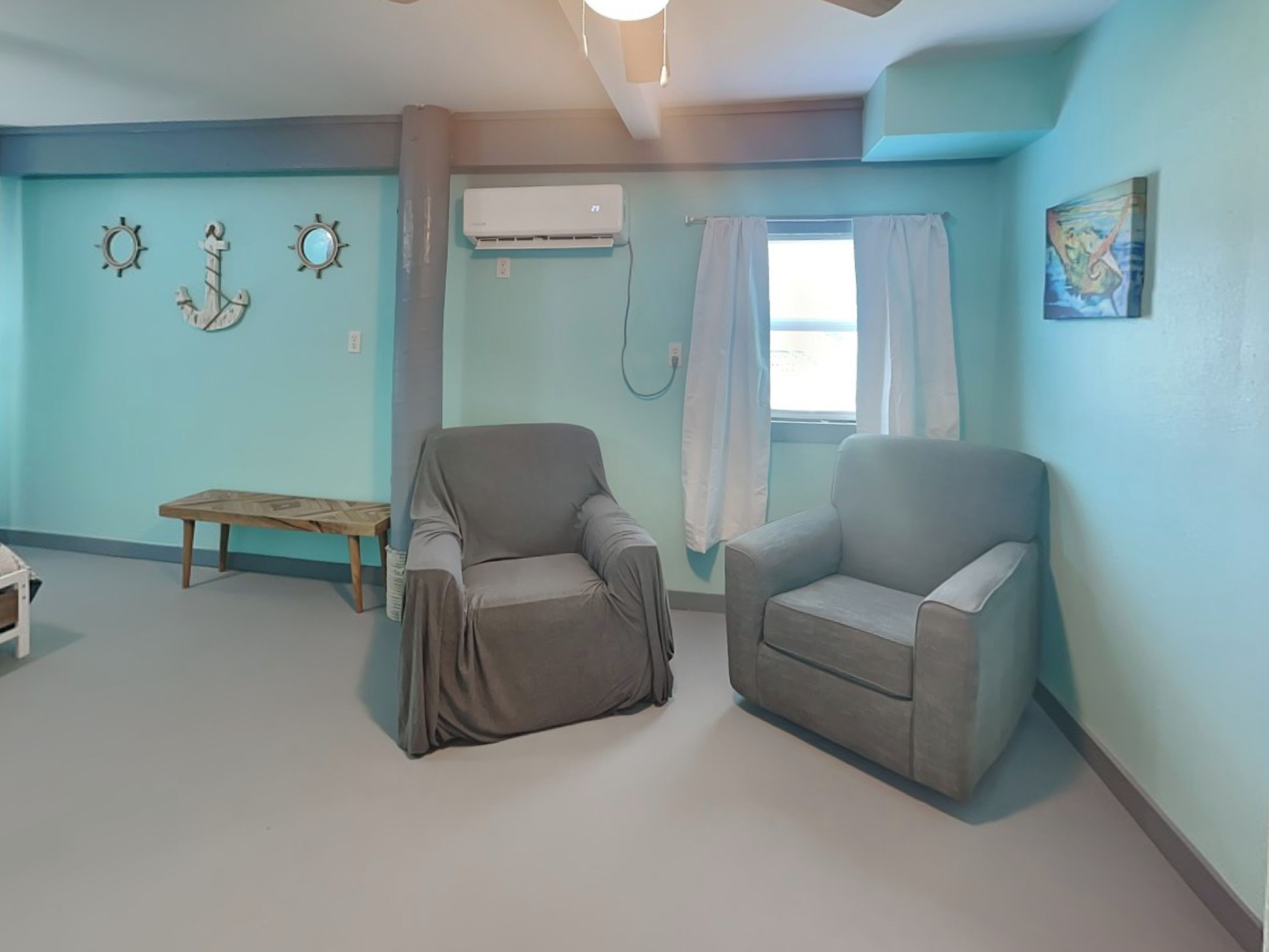 Een kamer met teal muren, twee grijze fauteuils, een houten bank en decoratie met een nautisch thema.