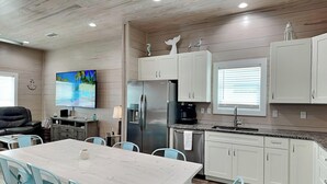 A modern kitchen with white cabinets, a stainless steel refrigerator, and a coffee maker. There is a dining area with a white table and turquoise chairs. - All Decked Out by Miss Kitty’s (Rockport)