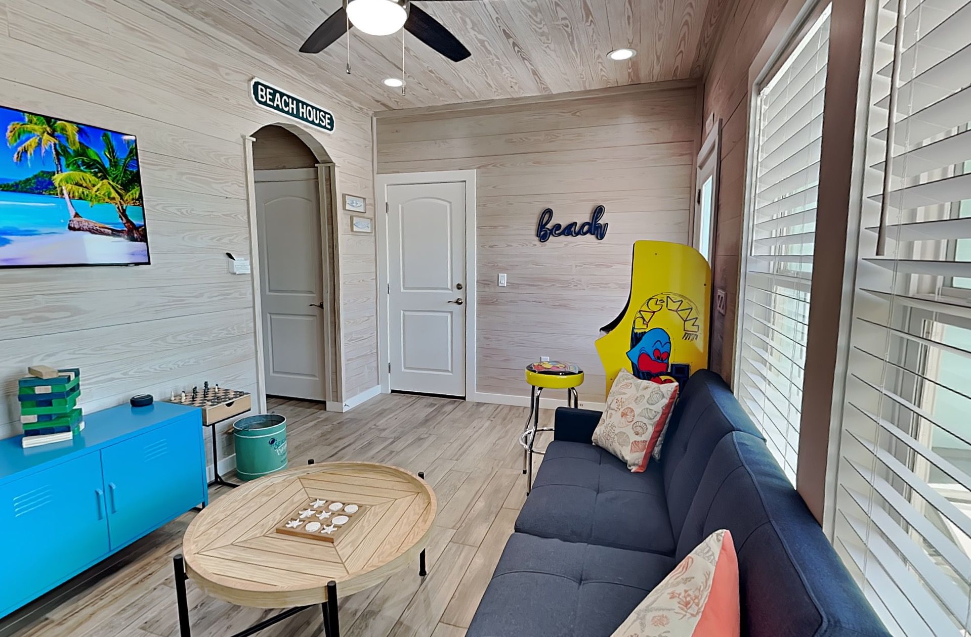 Ein Zimmer mit einem blauen Sofa, einem Holztisch, einem Fernseher, der eine Strandansicht zeigt, einer Tür mit der Aufschrift 'BEACH HOUSE' und einer Wand mit dem Wort 'strand'.