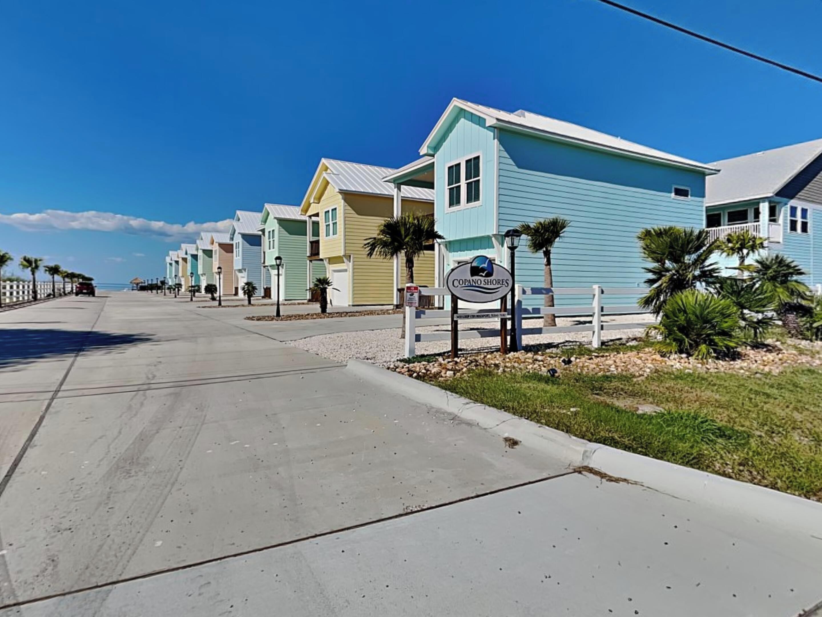 A coastal street lined with pastel-colored beach houses, a clear sky, and a sign for 'Copano Shores'.