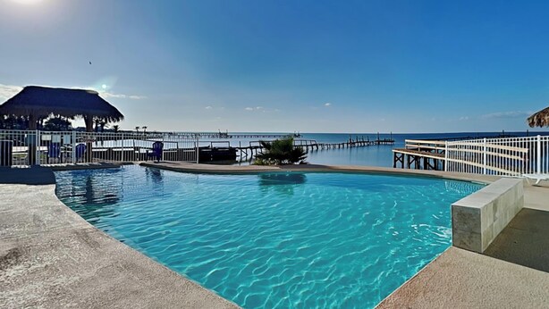 A clear blue pool with a wooden deck, a thatched-roof structure, and a pier extending into the water. - All Decked Out by Miss Kitty’s (Rockport)