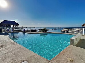 A clear blue pool with a wooden deck, a thatched-roof structure, and a pier extending into the water. - All Decked Out by Miss Kitty’s (Rockport)