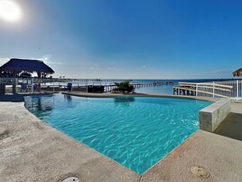 A clear blue pool with a wooden deck, a thatched-roof structure, and a pier extending into the water.