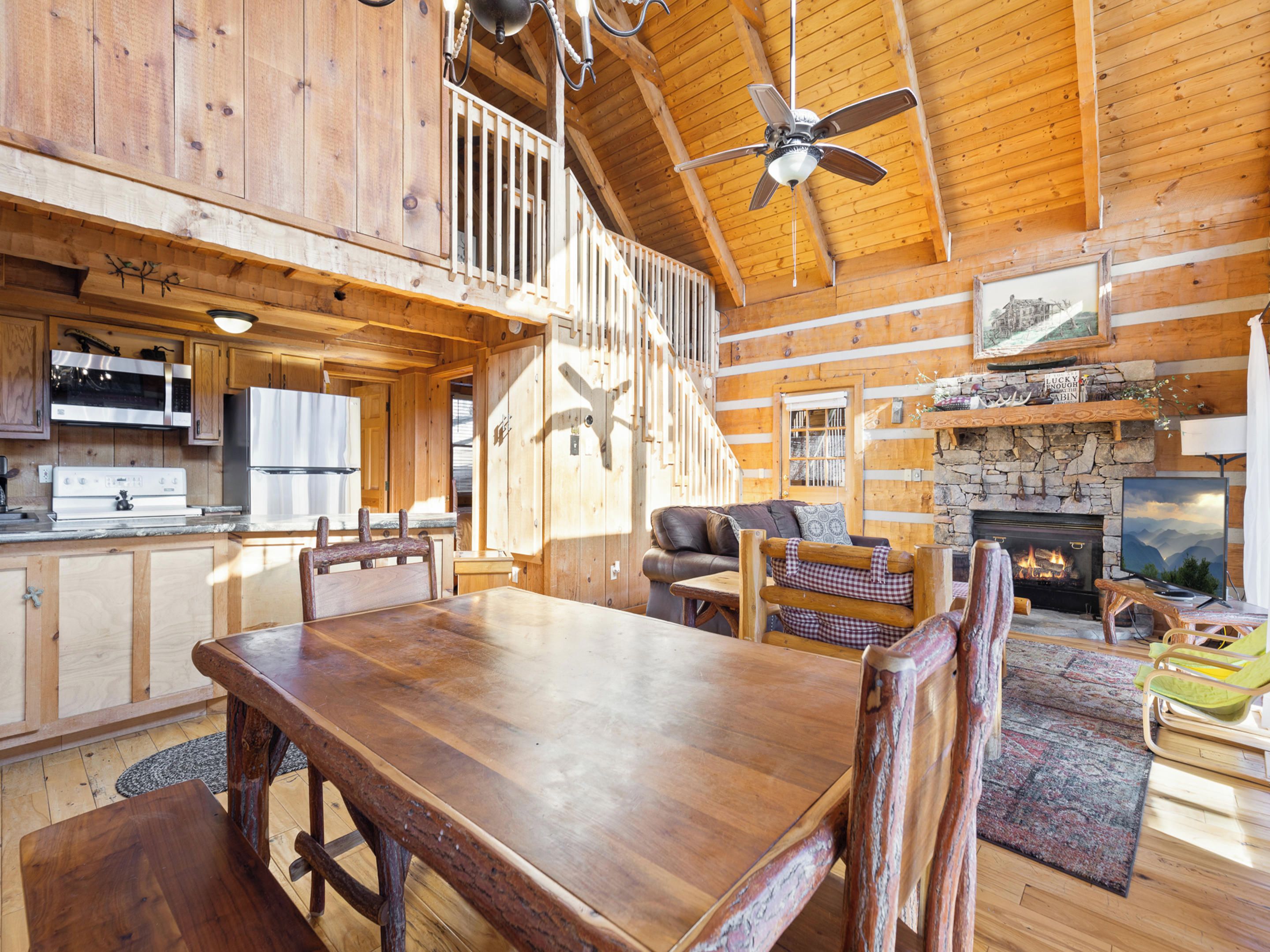 A rustic cabin interior with a dining area, kitchen, fireplace, and a loft bedroom.