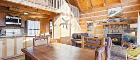 A rustic cabin interior with a dining area, kitchen, fireplace, and a loft bedroom.