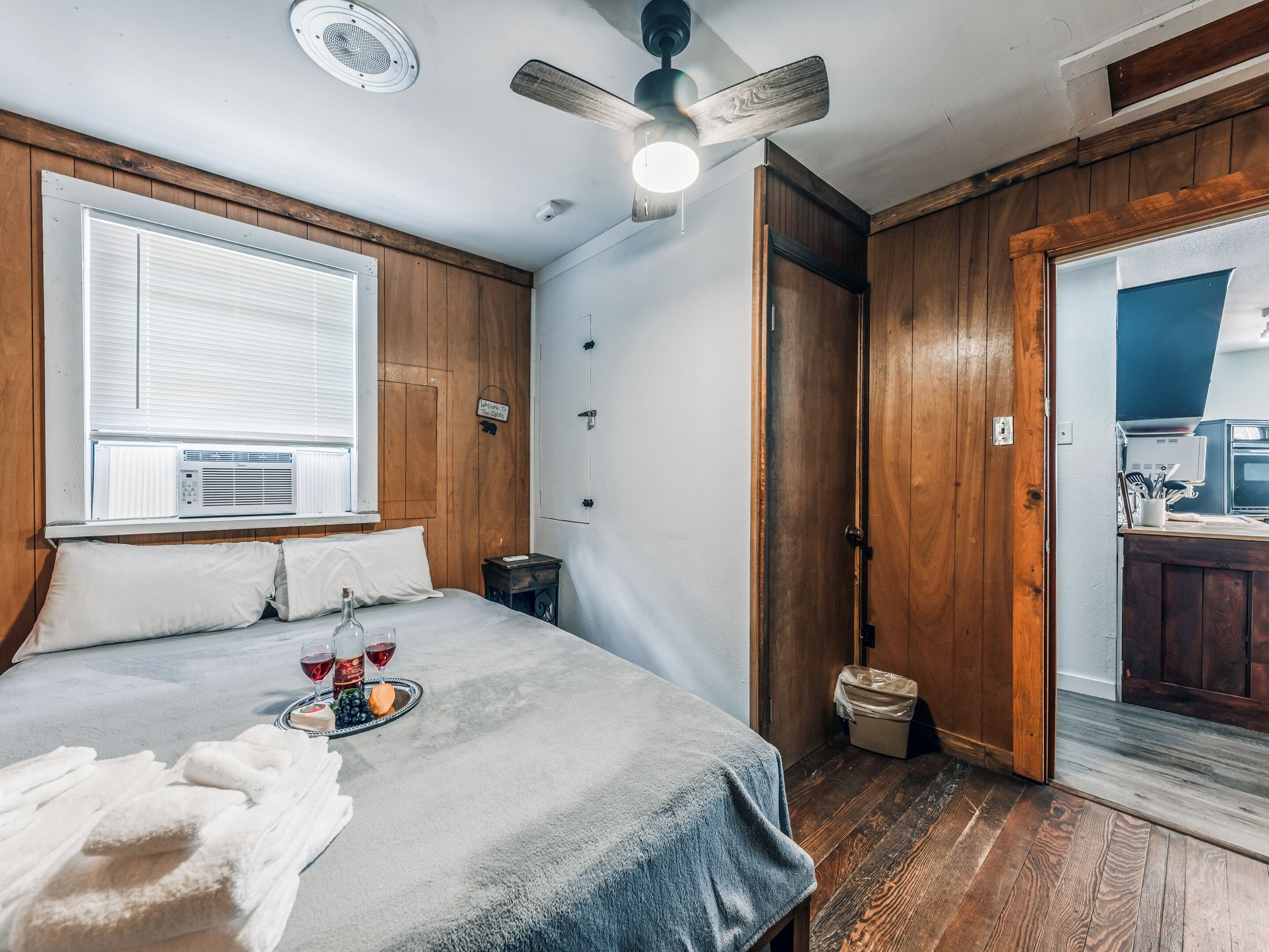 Un dormitorio con cama, ventilador de techo, ventana con persianas, mesita de noche y vista a la cocina a través de una puerta abierta.