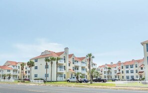 A row of multi-story residential buildings with balconies, parked cars, and palm trees. - Toes In The Water II by Silver Sands Vacation Rentals (Port Aransas)