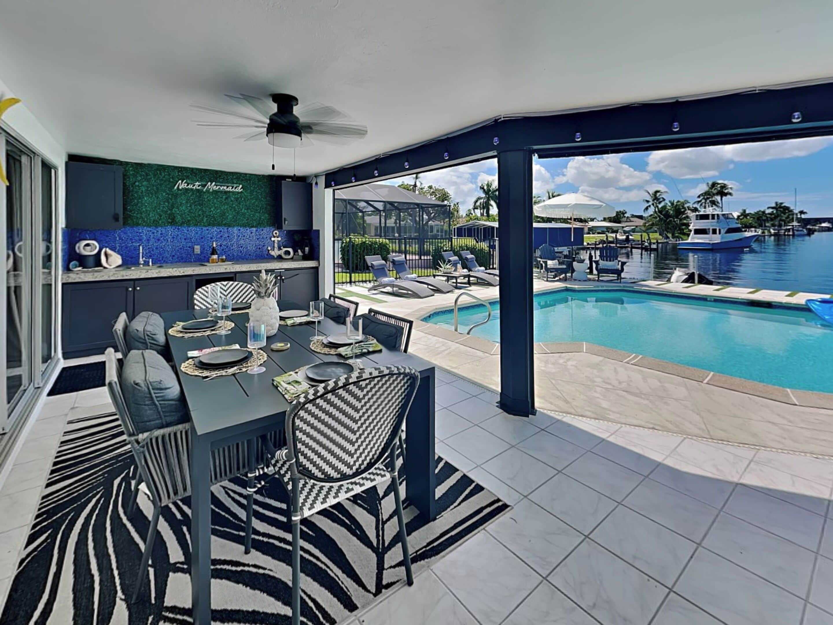 A modern outdoor dining area with a pool view, a ceiling fan, and a 'Nautic Mermaid' sign.