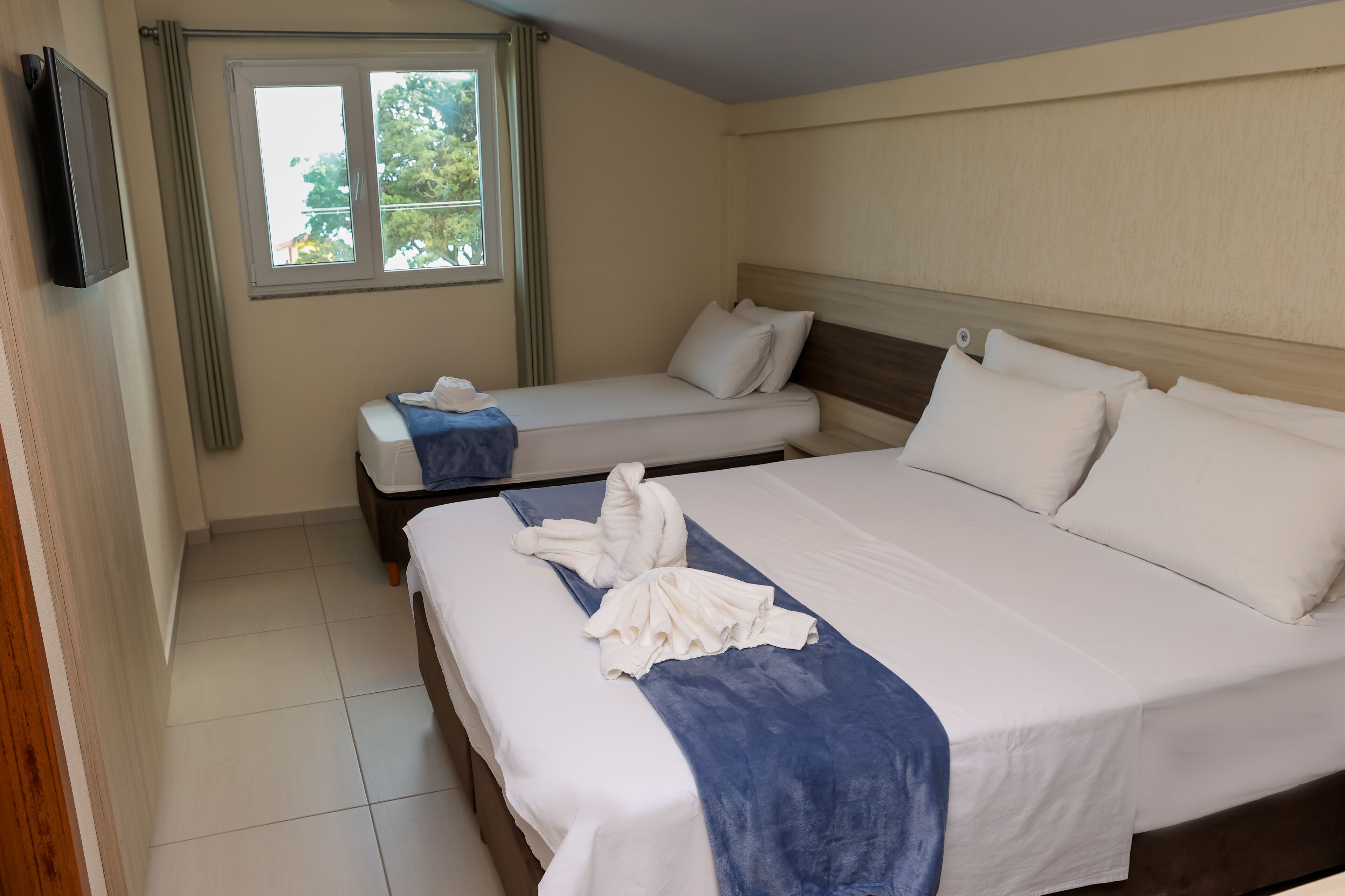 A hotel room with two beds, a TV, a window with curtains, and a view of greenery outside.