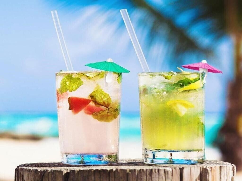 Two cocktails with straws and umbrellas on a wooden surface with a blurred beach and ocean in the background.