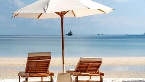 Dos sillones de madera bajo una sombrilla blanca en una playa con un mar tranquilo y un barco a lo lejos.