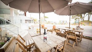 Un espacio de comedor al aire libre con mesas y sillas de madera, sombrillas y vista a un parque infantil.