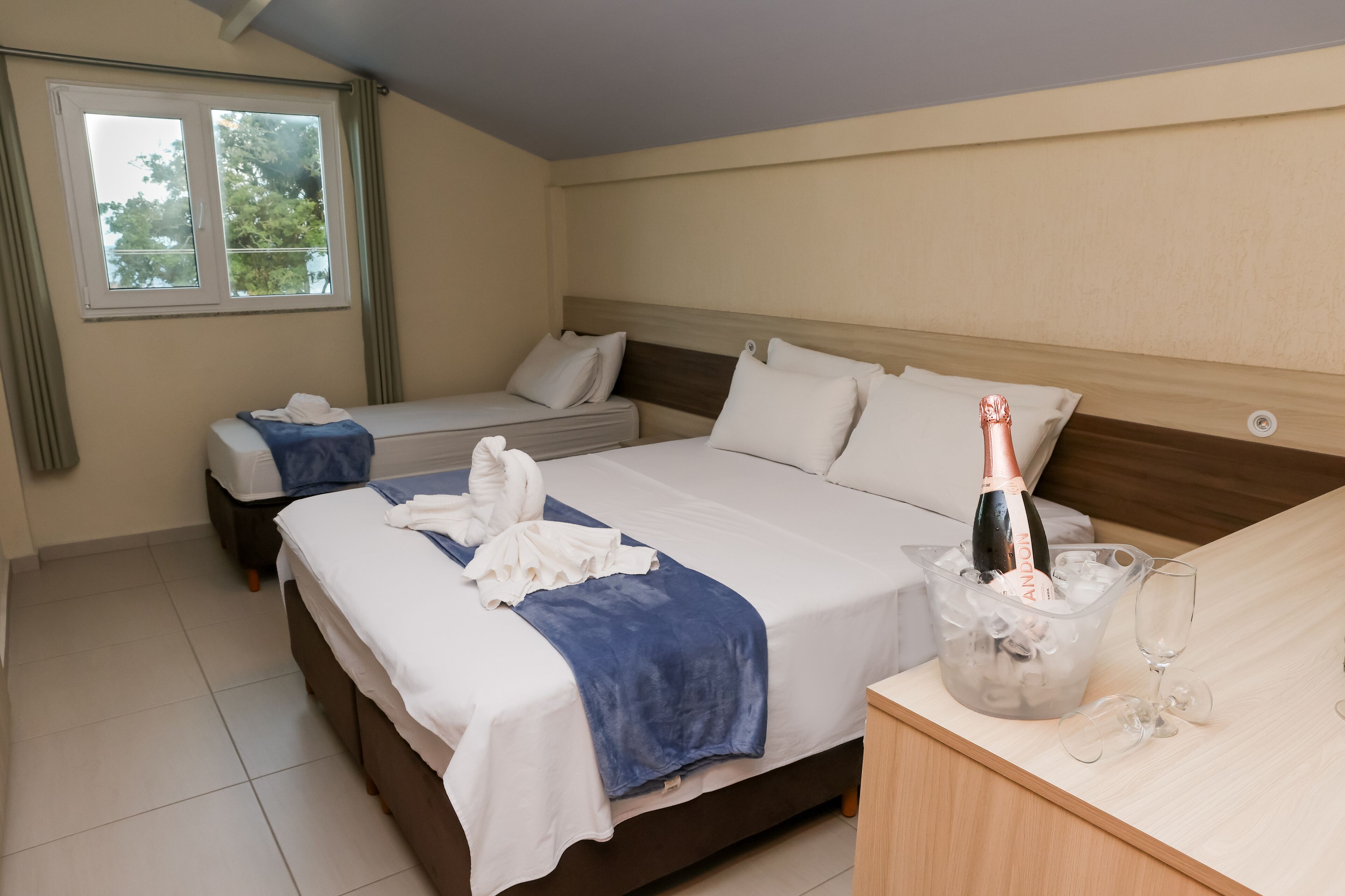 A hotel room with two beds, a minibar, and a window with a view of trees.