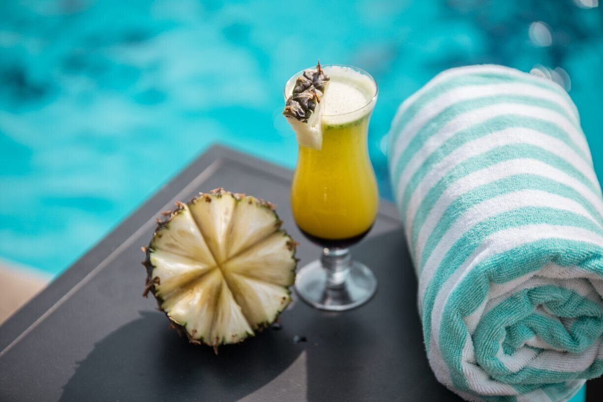 A tropical drink in a glass with a starfruit garnish, a halved pineapple, and a striped towel by a pool.