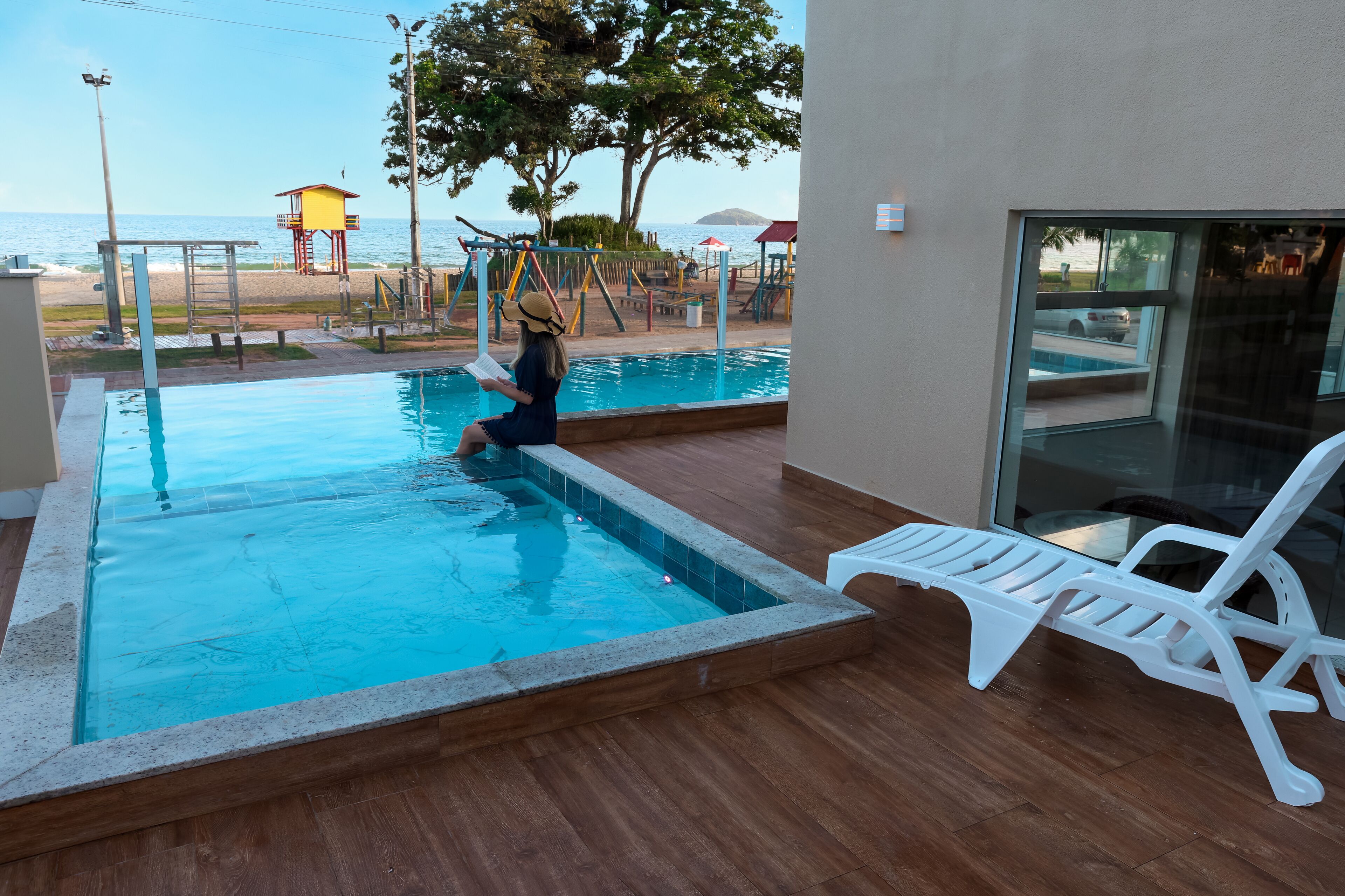Una piscina con una silla de salvavidas y un socorrista, un área de playa con una torre de vigilancia, y vista al mar.