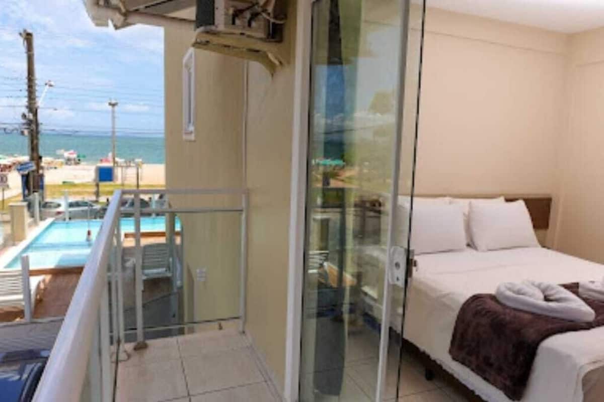A hotel room with a bed, a glass door leading to a balcony, and a view of the beach.