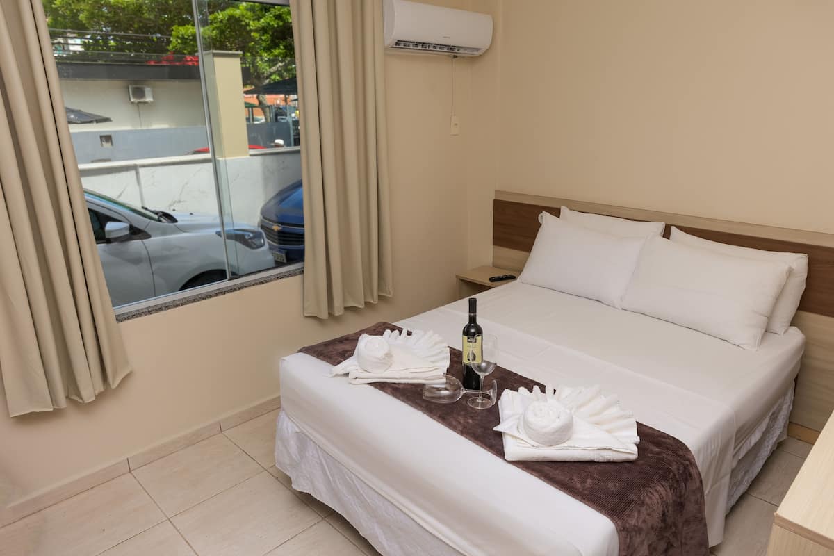A hotel room with a bed, a nightstand, a window with curtains, and a view of parked cars outside.