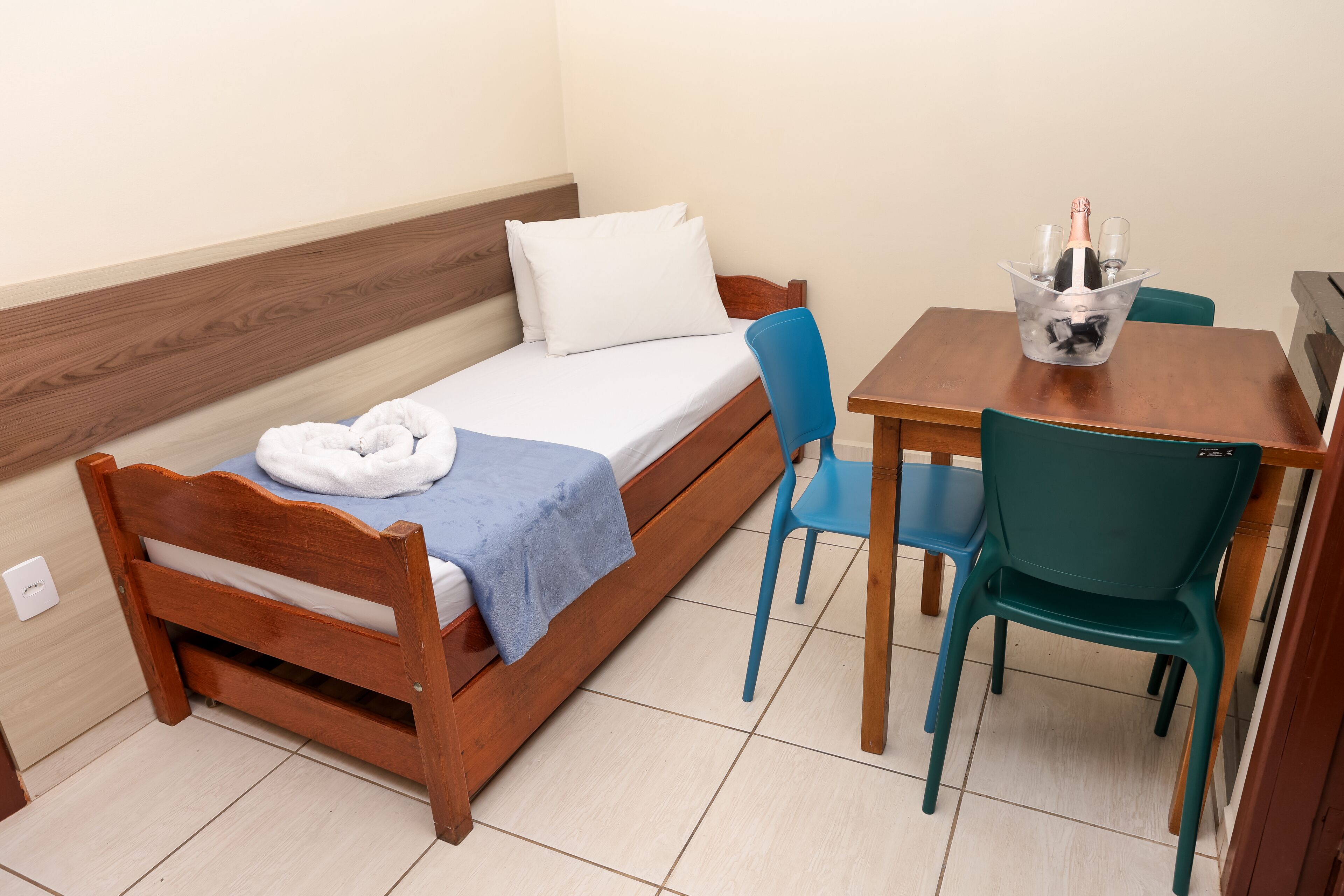 Una habitación pequeña con una cama individual, cabecera de madera, una toalla azul y una mesa con una botella y vasos.