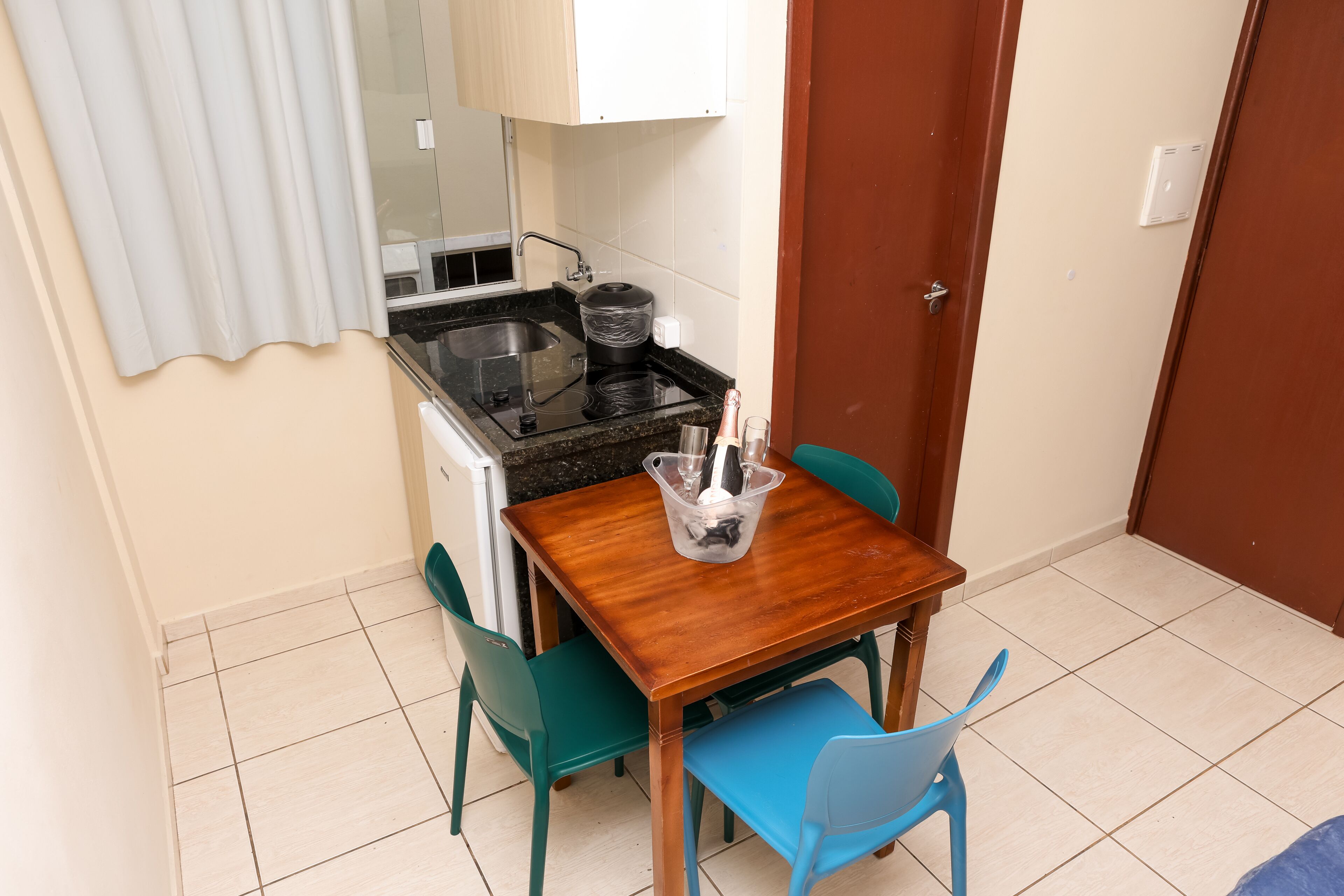 Una pequeña kitchenette con estufa, lavabo y armarios, una mesa de madera con sillas y una puerta a la derecha.