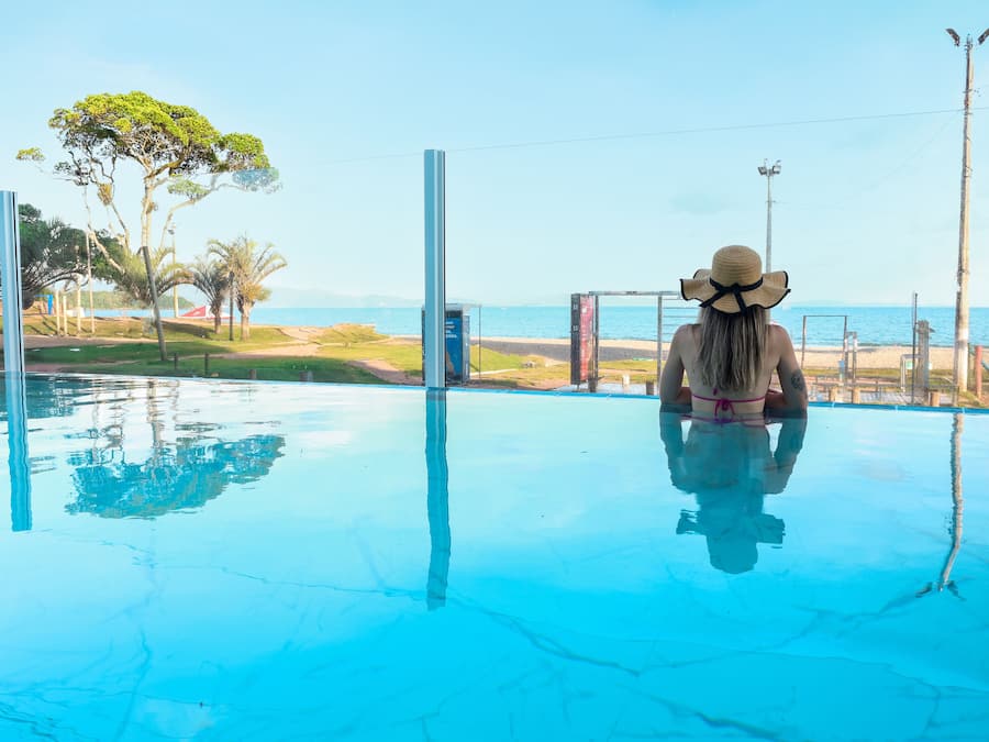 A person sitting by a clear blue pool with a view of the beach and palm trees.