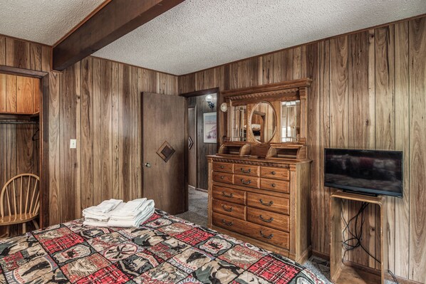 A bedroom with wooden walls, a bed with a patterned quilt, a wooden dresser with drawers, a mirror, and a television on a stand. - Knott Inn by Ruidoso Vacation Rentals (Ruidoso)