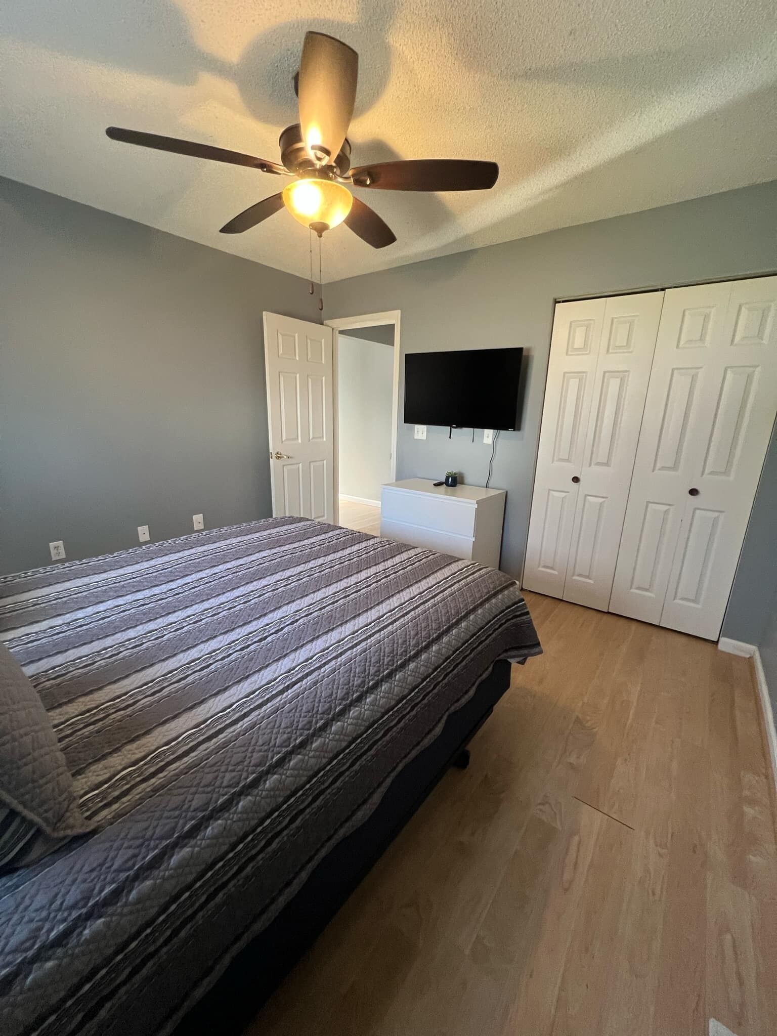 A bedroom with a bed, a ceiling fan, a TV mounted on the wall, and a wardrobe.
