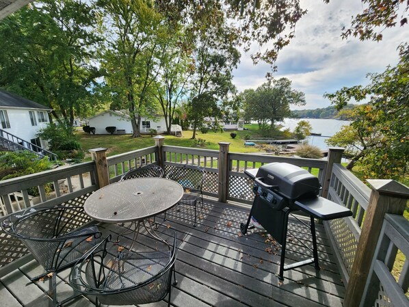 A deck with a round table and chairs, a grill, and a view of a lake and houses in the background. - Relaxing lake front home with beautiful view on the lake, sleeps 7.  (Creal Springs)