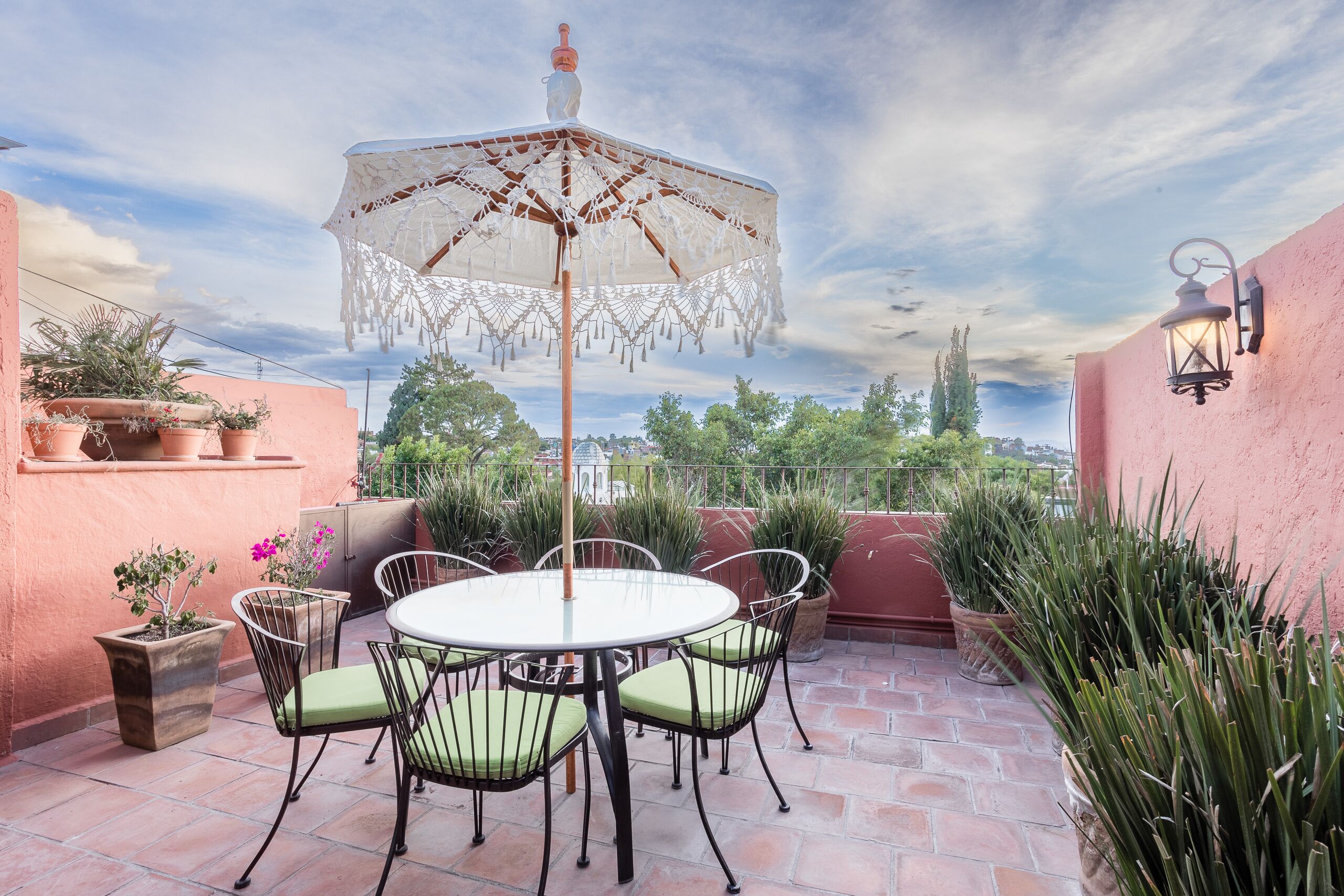 Un patio con una mesa redonda, sillas, un paraguas decorativo, plantas en macetas y vistas de la ciudad.
