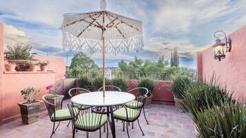 Un patio con una mesa redonda, sillas, un paraguas decorativo, plantas en macetas y vistas de la ciudad.