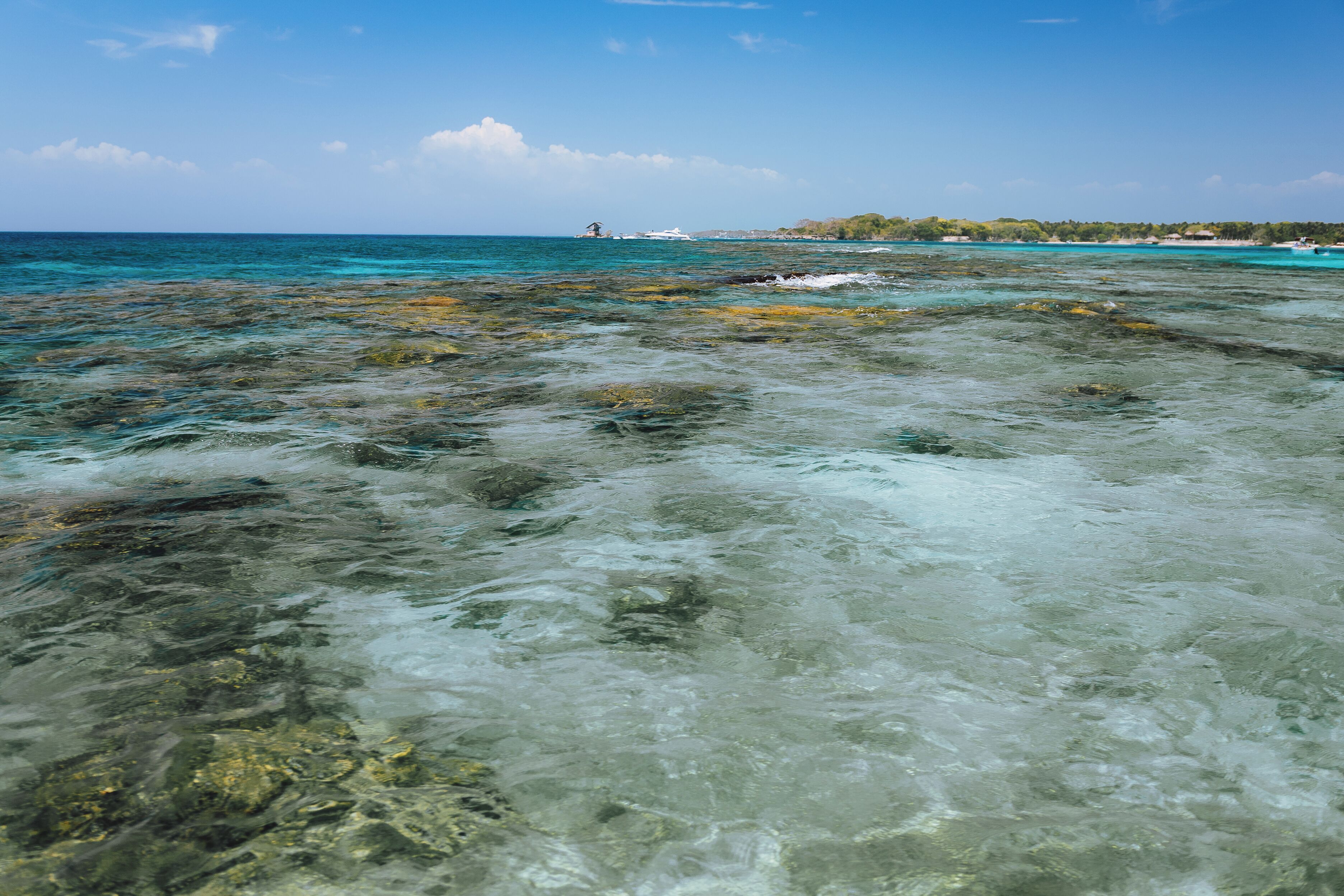 A clear view of a rocky shoreline with shallow water, a distant boat, and a clear sky.