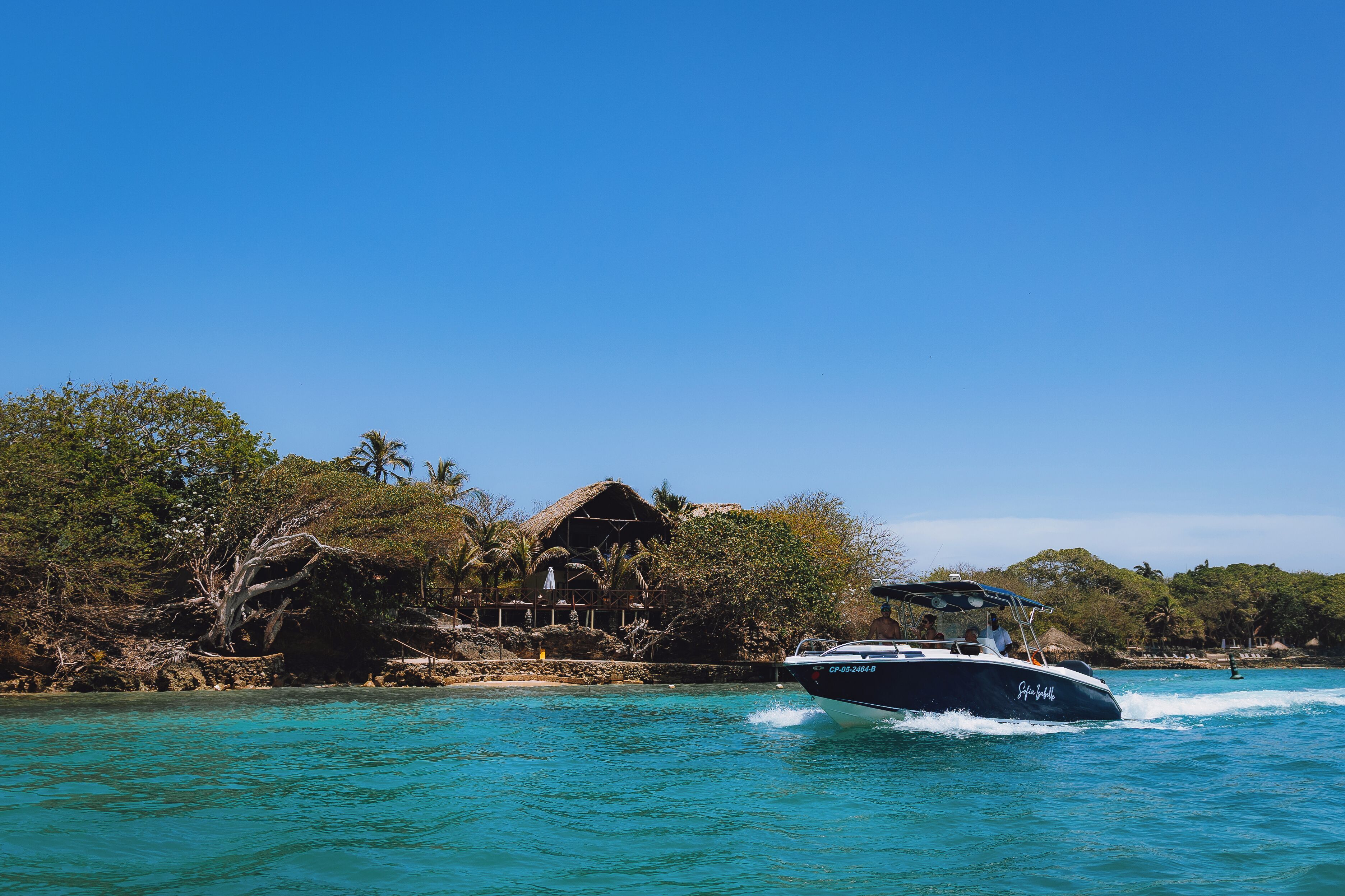 A boat moving through turquoise waters with a thatched-roof structure and lush greenery in the background.