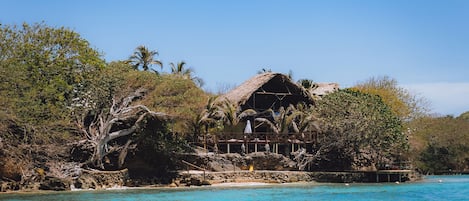 A beachfront property with a thatched-roof structure, surrounded by dense tropical vegetation and a clear blue sea.