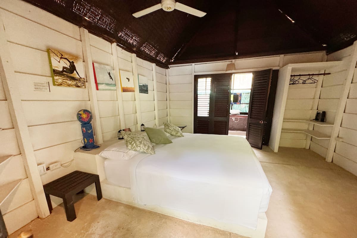 A bedroom with a large bed, a bench, a wall-mounted shelf, and a door leading to an outdoor area.