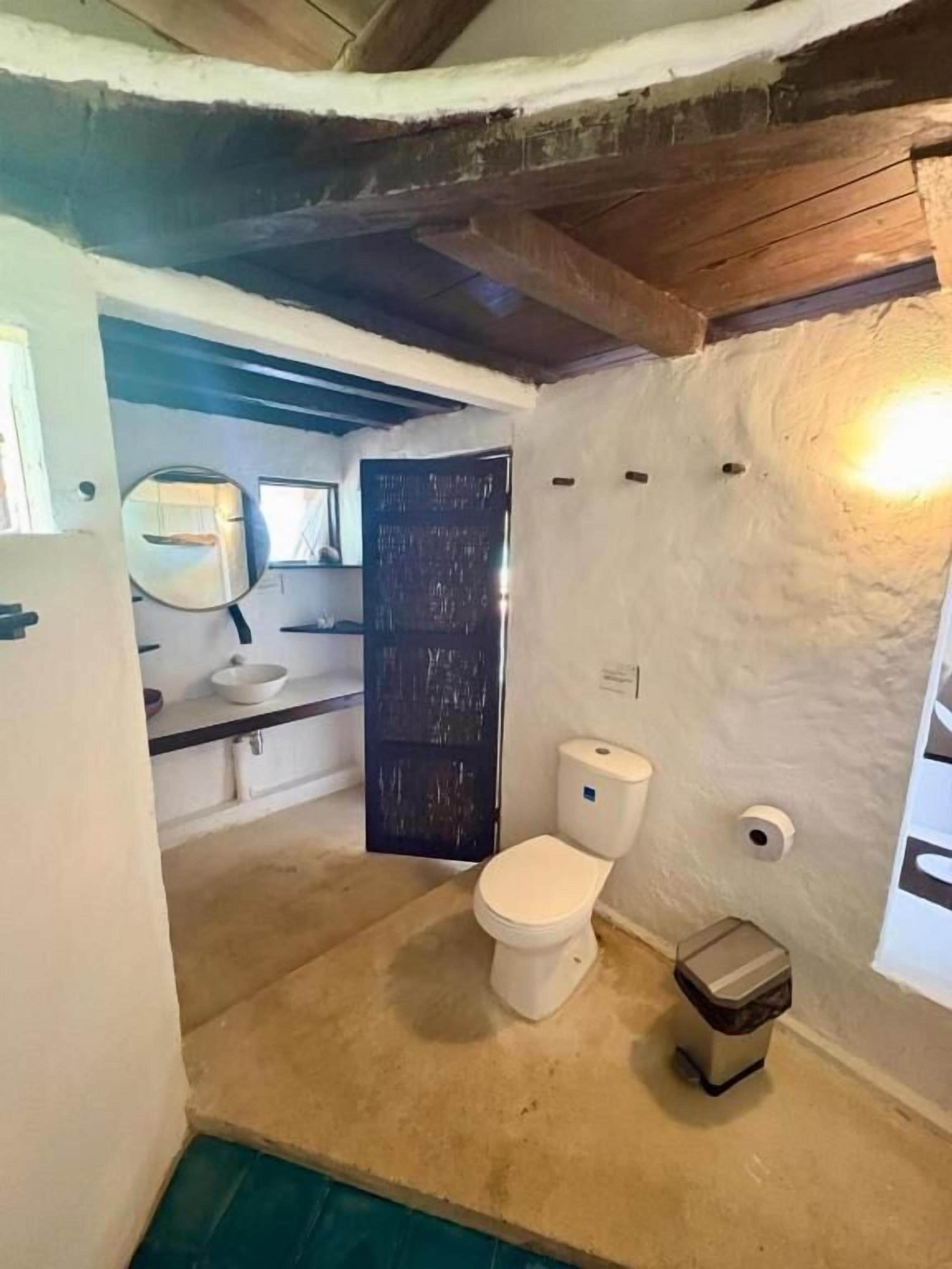 A small, rustic bathroom with a white toilet, a sink, and a mirror. The walls are textured, and there's a wooden beam ceiling.