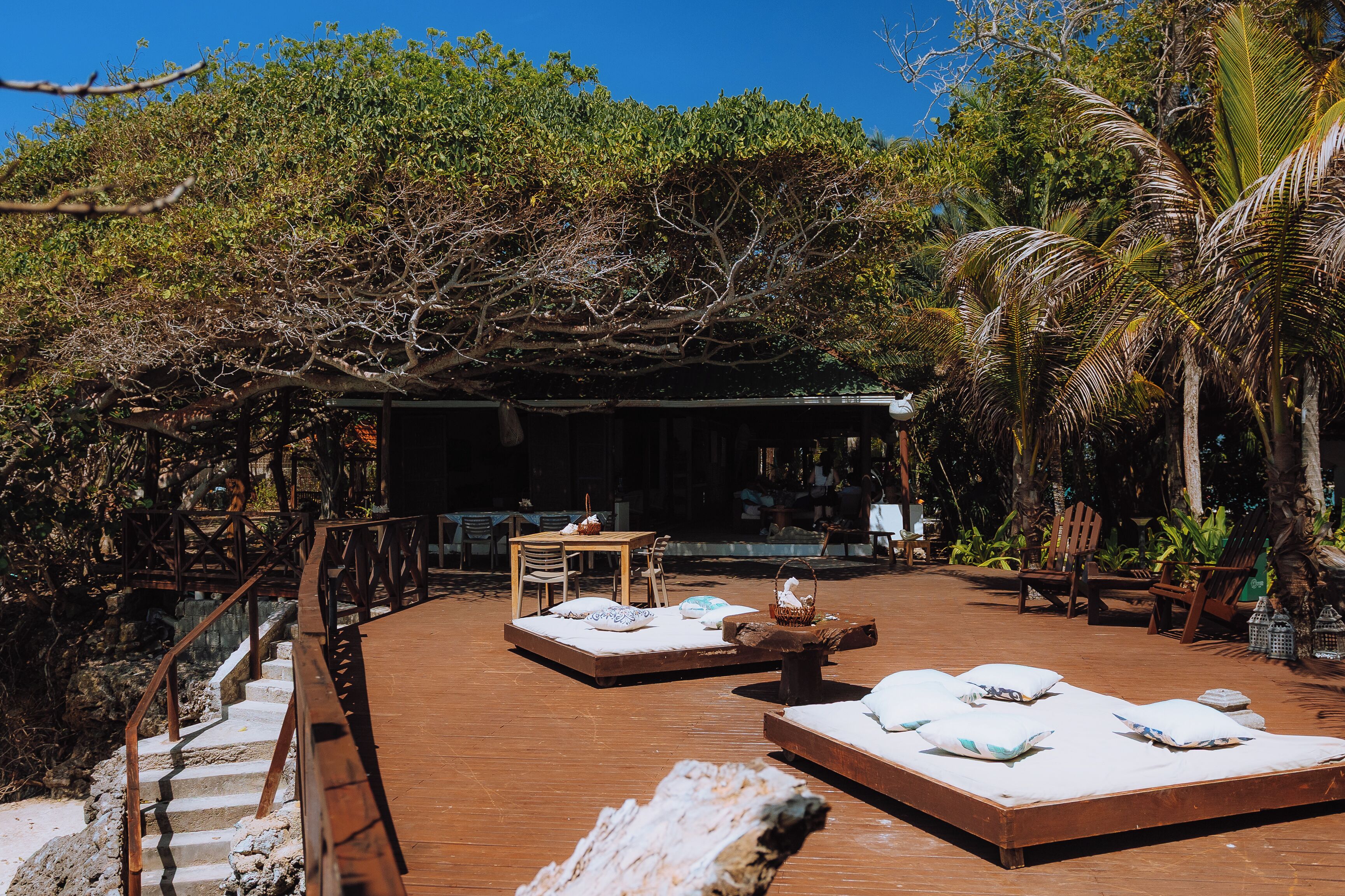 A wooden deck with a bed and chairs, surrounded by trees and a covered area.