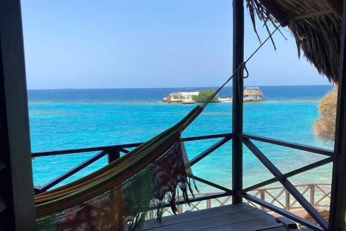 A wooden deck with a hammock and a wooden chair, overlooking turquoise waters and a small island.