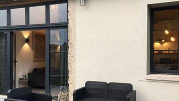 Een terras buiten met rieten meubilair, een betegelde vloer en een gebouw met betegeld dak en grote ramen.