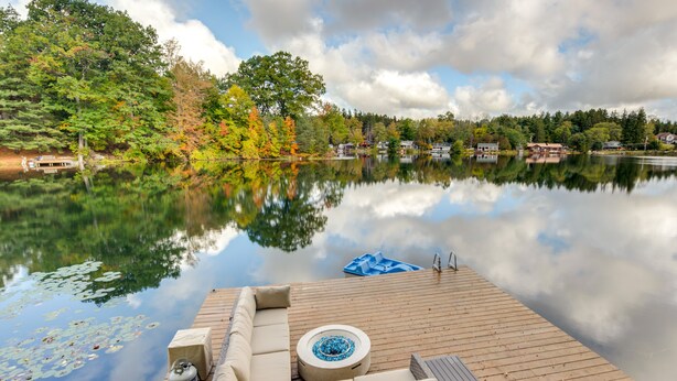 Una terraza de madera con un sofá y un hogar, con vista a un lago tranquilo que refleja árboles y casas.